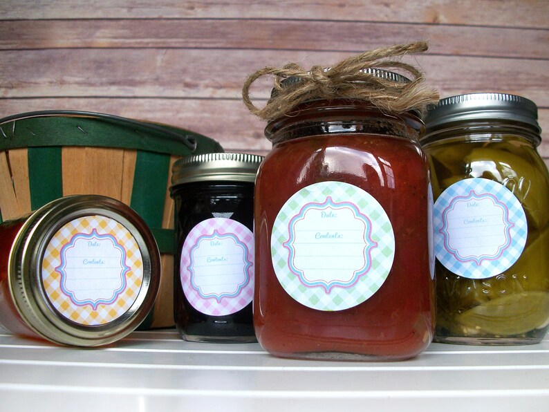 May include: Four glass jars with labels on them. The labels are round and have a gingham pattern. The jars are filled with different types of preserves. The jars are sitting on a white surface.