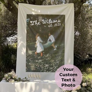 May include: A white fabric backdrop with a printed photo of a couple, the text "The Wilsons est. 2025". A white table is set with champagne flutes and floral arrangements, perfect for a wedding or event.