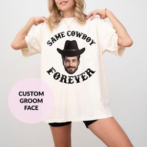 May include: A white t-shirt with the text "Same Cowboy Forever" in black lettering. A black cowboy hat with a face inside is in the centre of the text. The text is in a distressed font.