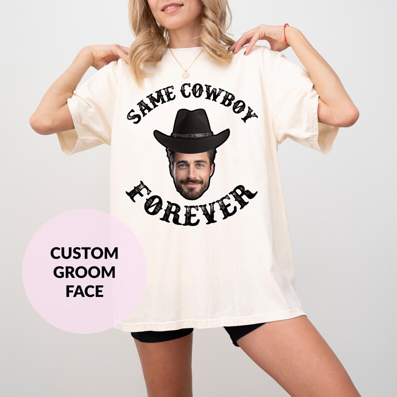 May include: A white t-shirt with the text "Same Cowboy Forever" in black lettering. A black cowboy hat with a face inside is in the centre of the text. The text is in a distressed font.