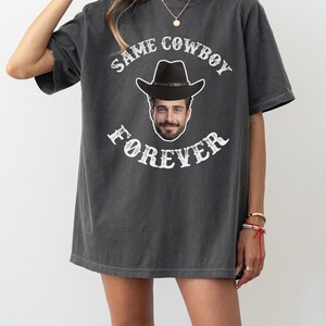May include: A charcoal grey t-shirt with a white graphic print of a cowboy hat and a man's face. The text on the shirt reads "Same Cowboy Forever".