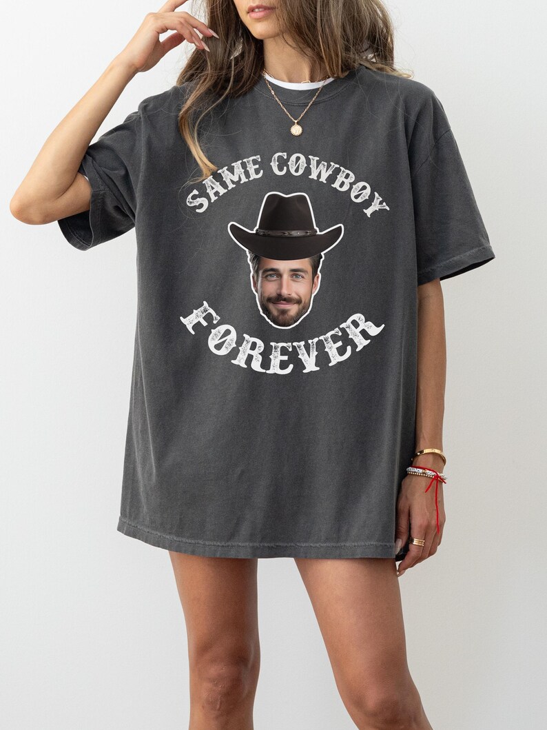 May include: A charcoal grey t-shirt with a white graphic print of a cowboy hat and a man's face. The text on the shirt reads "Same Cowboy Forever".
