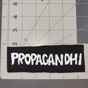Può includere: Una toppa rettangolare nera con la scritta "PROPAGANDHI" in lettere bianche e in grassetto. La toppa è posizionata su un tappetino da taglio grigio con linee a griglia e un righello.
