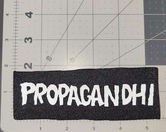 May include: A rectangular black patch with the word "PROPAGANDHI" in bold white letters. The patch is placed on a gray cutting mat with a ruler and grid lines.