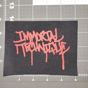 Puede incluir: Parche rectangular negro con la inscripción "IMMORTAL TECHNIQUE" en rojo, con un estilo graffiti. El parche está sobre una esterilla de corte gris con marcas en centímetros.