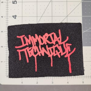 May include: A black rectangular patch with the words "IMMORTAL TECHNIQUE" in bright red, stylized graffiti-style lettering. The patch is placed on a gray and white grid background.