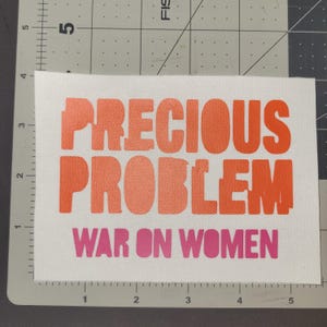 Könnte beinhalten: Ein weißes Stoffabzeichen mit den Worten "PRECIOUS PROBLEM" in Orange und "WAR ON WOMEN" in Pink. Der Text ist in einer fetten, serifenlosen Schriftart.