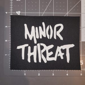 Peut inclure: Écusson rectangulaire noir avec l'inscription "MINOR THREAT" en lettres blanches en gras, de style pochoir. L'écusson est posé sur une surface grise avec un quadrillage.