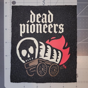 May include: Black square patch with the words "dead pioneers" in white gothic font. Below the text is a skull, a covered wagon, and a red flame design. The wagon wheels and skull are beige.
