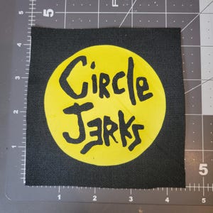 May include: Square black patch featuring a yellow circle with the words "Circle Jerks" in black, stylized lettering. The patch measures approximately 10 cm square. Ideal for jackets or bags.