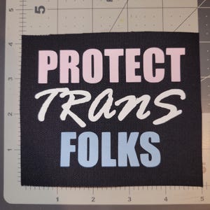 May include: Black fabric patch with the words "PROTECT TRANS FOLKS" in pink, white, and light blue lettering. The patch is rectangular and sits on a gray and white grid.