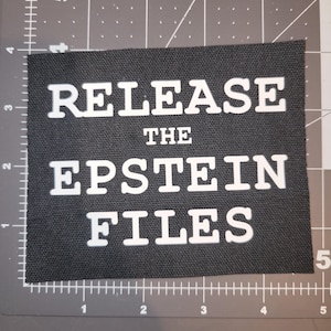 Peut inclure: Un écusson de tissu noir rectangulaire avec l'inscription blanche "RELEASE THE EPSTEIN FILES". Les lettres sont en caractères gras et sans empattement. Le patch est probablement en tissu tissé.
