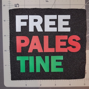 May include: Black square patch with the words "FREE PALESTINE" in white, red, and green block letters. The patch is on a gray surface with a ruler.
