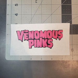 Puede incluir: Un parche rectangular de tela blanca con el texto THE VENOMOUS PINKS en una fuente rosa estilizada con contornos negros y un efecto de goteo. El parche está colocado sobre una base de corte con marcas de pulgadas visibles en el borde.