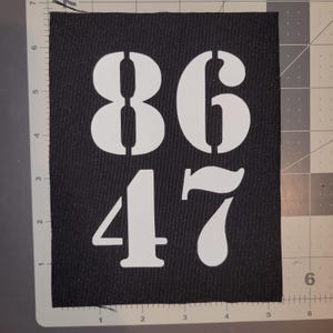 Puede incluir: Parche rectangular negro con los números 86 y 47 en fuente de plantilla blanca. El parche está sobre una esterilla de corte gris con una regla en el borde.