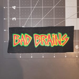 Pode incluir: Patch retangular preto com as palavras "BAD BRAINS" em letras vermelhas e amarelas. O patch tem uma borda verde e está em um fundo preto. Adequado para roupas ou acessórios.