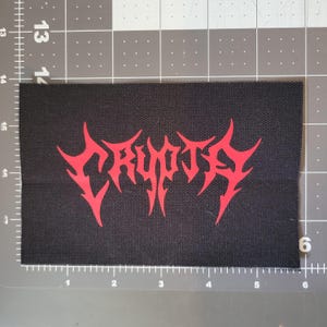 Puede incluir: Parche rectangular negro con la palabra "CRYPT" en rojo, con letras puntiagudas. El parche está hecho de tela tejida, probablemente para planchar o coser en ropa o accesorios. Mide aproximadamente 10 cm x 6 cm.
