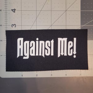 May include: A rectangular black fabric patch featuring the white text Against Me! in a stylized font. The patch is positioned on a cutting mat with a visible grid, showing the patch measures approximately 5.5 inches in width and 2.5 inches in height.
