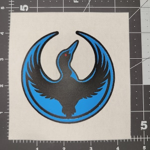May include: A square patch with a black bird emblem inside a blue crescent. The bird has a red eye. The design is set against a white canvas background.