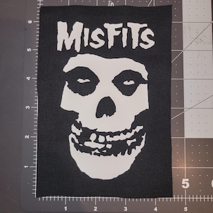 May include: Black rectangular patch featuring the word "Misfits" in white above a white skull graphic. The patch is made of a woven fabric. The skull has a wide grin and is a recognizable symbol.