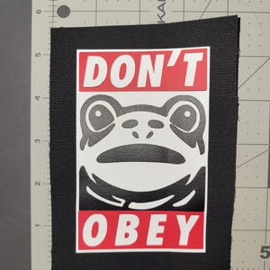 Può includere: Toppa rettangolare nera con un volto di rana bianco e le parole "DON'T OBEY" in bianco su sfondo rosso. La toppa misura circa 10 cm per 7,6 cm.