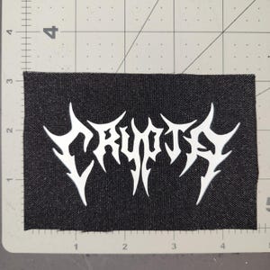 Peut inclure: Écusson en tissu noir avec le mot "CRYPTA" blanc dans une police gothique pointue. L'écusson est rectangulaire et semble être fait d'un matériau tissé. L'écusson est sur une surface grise avec des mesures en centimètres.