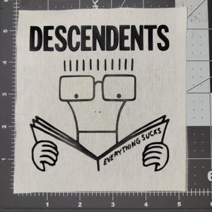Könnte beinhalten: Quadratischer, cremefarbener Aufnäher mit dem Wort "DESCENDENTS" in schwarzen Buchstaben. Darunter eine Comicfigur mit Brille, die ein Buch mit der Aufschrift "EVERYTHING SUCKS" liest.