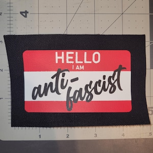 Może przedstawiać: Prostokątna czarna naszywka z tkaniny z grafiką czerwono-białego identyfikatora. Tekst HELLO I AM jest napisany białymi literami, a słowo anti-fascist jest nadrukowane czarną kursywą na środkowym białym poziomym pasku naszywki.