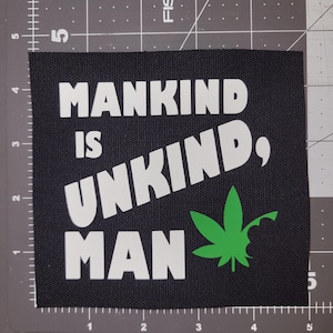 May include: Black square patch with the text "MANKIND IS UNKIND, MAN" in white, with a green cannabis leaf graphic. The patch is on a gray and black grid.
