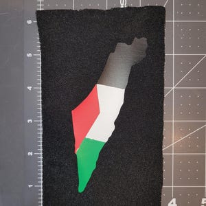 May include: A rectangular black patch with a map of a country in red, white, green, and black. The top of the map has a gradient effect. The patch measures approximately 15 cm high and 10 cm wide.