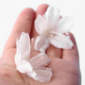 May include: Two delicate, translucent white artificial flowers with five rounded petals each and thin, prominent stamens extending from the center. The blossoms feature a soft, semi-sheer texture and are positioned to show their layered floral structure.