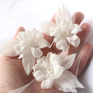 Pode incluir: Três enfeites de flores de tecido branco com pétalas delicadas e um centro texturizado. As flores são dispostas sobre um tecido branco transparente, criando um visual suave e etéreo. Ideal para projetos de casamento ou artesanato.
