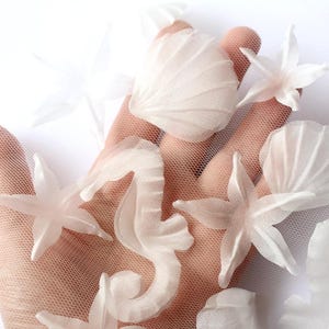 Puede incluir: Una mano sosteniendo una variedad de decoraciones marinas de tela blanca, incluyendo una concha, una estrella de mar y un caballito de mar.