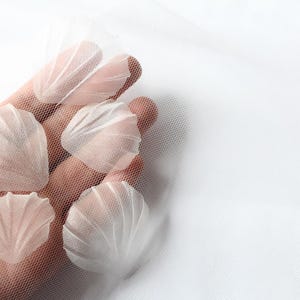 Könnte beinhalten: Fünf zarte weiße Stoffblumenblätter, die Muscheln ähneln, werden in einer Hand vor einem weißen Tüllhintergrund gehalten.