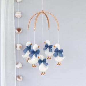 May include: A nursery mobile with six white geese, each adorned with a blue bow, hanging from a wooden frame. The geese have orange beaks and feet. The background is light grey, with pom-poms hanging on the left side.