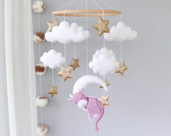 Op de afbeelding: Baby mobile met een houten ring, witte vilten wolken, gouden glittersterren en een slapende paarse draak op een witte maan. Een hangende decoratie voor de kinderkamer.