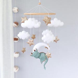 May include: A baby mobile featuring a wooden ring with hanging white clouds, gold glitter stars, and a sleeping teal dragon on a white crescent moon. The mobile is suspended by white strings.