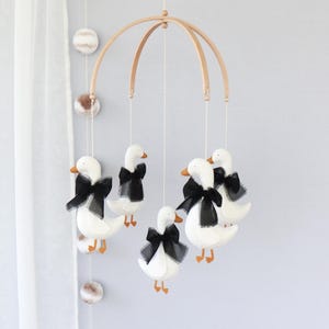 Op de afbeelding: Een baby mobile met witte ganzenfiguren, oranje snavels en poten, en zwarte strikken. De ganzen hangen aan een houten boog, een charmante decoratie voor de kinderkamer.