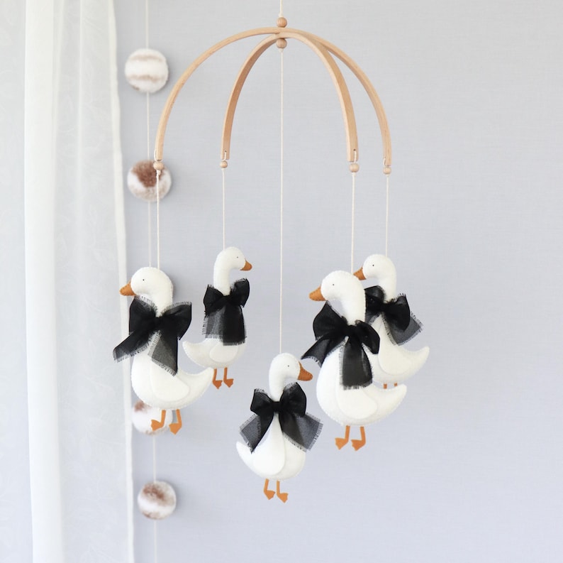 Op de afbeelding: Een baby mobile met witte ganzenfiguren, oranje snavels en poten, en zwarte strikken. De ganzen hangen aan een houten boog, een charmante decoratie voor de kinderkamer.