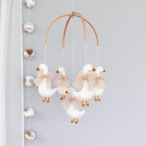 May include: A baby mobile featuring six white geese with orange beaks and feet, each adorned with a light pink bow. The geese are suspended from a wooden arched frame. The background is a light gray wall with decorative pom-poms.