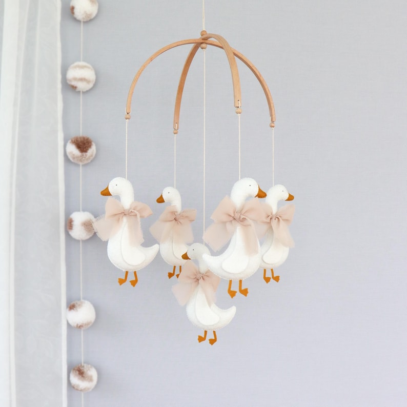 May include: A baby mobile featuring six white geese with orange beaks and feet, each adorned with a light pink bow. The geese are suspended from a wooden arched frame. The background is a light gray wall with decorative pom-poms.