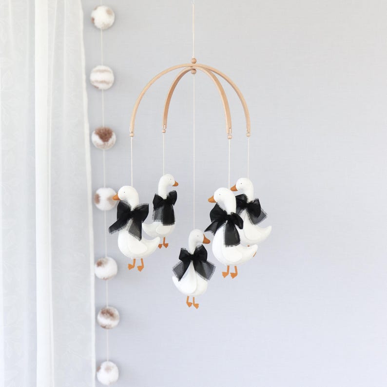 Op de afbeelding: Een baby mobile met vijf witte ganzen met zwarte strikken en oranje snavels. De ganzen hangen aan een houten boog. De mobile hangt voor een lichtgrijze muur, met pompons aan de linkerkant.