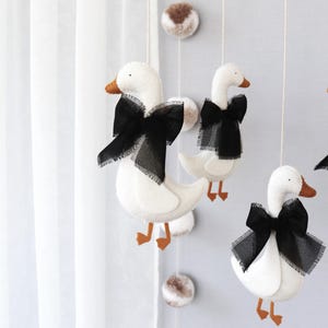 Op de afbeelding: Een decoratieve mobiel met witte vilten ganzen, zwarte strikken en oranje snavels en poten. De ganzen hangen aan touwtjes met pompons, wat een charmante kinderkamerdecoratie cre&euml;ert.