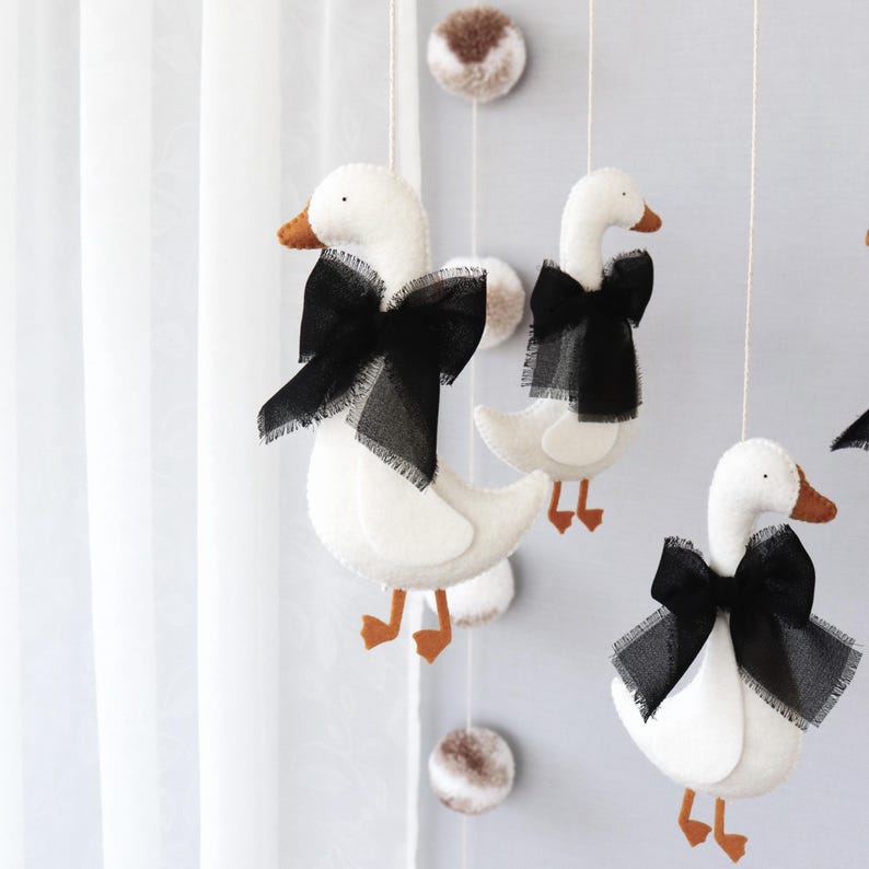 Op de afbeelding: Een decoratieve mobiel met witte vilten ganzen, zwarte strikken en oranje snavels en poten. De ganzen hangen aan touwtjes met pompons, wat een charmante kinderkamerdecoratie cre&euml;ert.