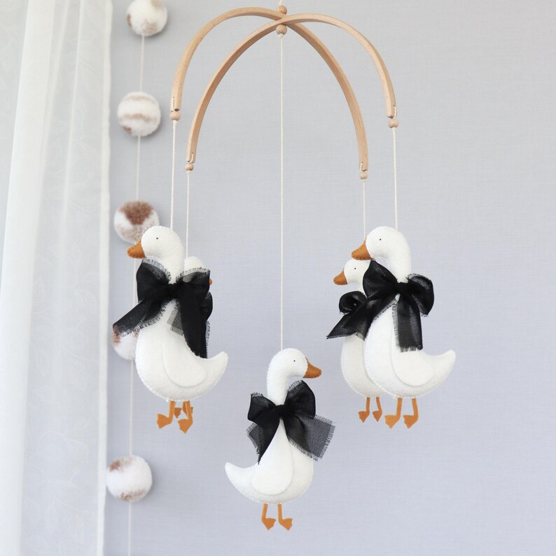 Op de afbeelding: Een baby mobile met witte vilten ganzen, oranje snavels en poten, en zwarte satijnen strikken. De ganzen hangen aan een natuurlijk houten frame met witte touwtjes. Decoratieve pompons zijn ook opgehangen.