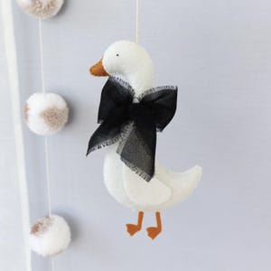 Op de afbeelding: Een witte vilten mobiel met een gans met een zwarte strik en oranje snavel en poten. De gans hangt aan een wit touwtje, met pompons op de achtergrond. Handgemaakte kinderkamerdecoratie.