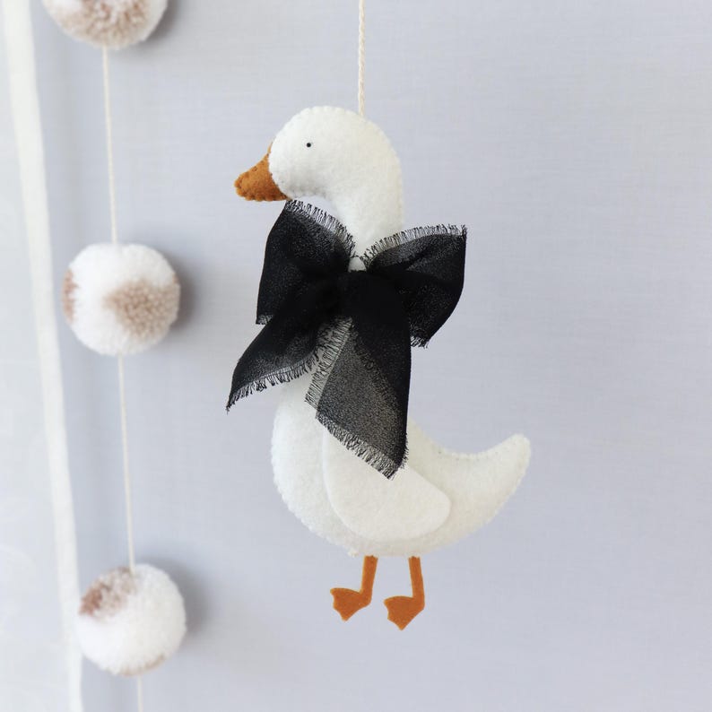 Op de afbeelding: Een witte vilten mobiel met een gans met een zwarte strik en oranje snavel en poten. De gans hangt aan een wit touwtje, met pompons op de achtergrond. Handgemaakte kinderkamerdecoratie.