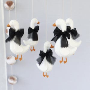 Op de afbeelding: Een mobiel met vijf witte vilten ganzen met oranje snavels en poten, elk versierd met een zwarte strik. De ganzen hangen aan witte touwtjes, met decoratieve pompons in de buurt. De achtergrond is zachtgrijs.