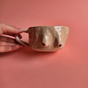 May include: A beige ceramic mug with a glossy finish and a textured, uneven surface. The mug features two raised, rounded protrusions on the side, each topped with a small, distinct dark red painted dot. It has a curved handle on the left side.
