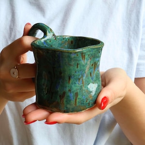 May include: A handmade ceramic mug with an irregular, organic shape and a glossy glaze in shades of deep forest green, teal, and brown. The mug features a small, curved handle at the top rim and a textured, uneven surface throughout the exterior.